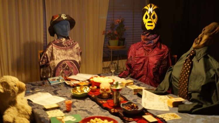 Four characters sitting around a table: a teddy bear, a blue-faced woman, a woman in a wrestling mask and wood-sculpture faced man