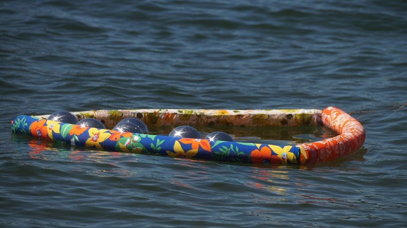Three pool noodles covered in patterned spandex and tied together in a triangle, creating a cage for six reflection balls floating on a lake.